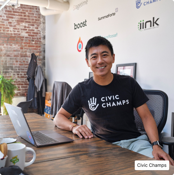 CIVIC CHAMPS – MAN SITTING AT OFFICE TABLE/LAPTOP SMILING AT CAMERA